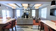 This single-floor space presents a well-appointed lounge or common area designed with both comfort and visual appeal in mind. The room's most prominent feature is a floor-to-ceiling accent wall surrounding a sleek fireplace, clad in mixed-tone reclaimed-style wood planking that creates a striking focal point. Flanking the fireplace, two generously sized windows allow natural light to filter into the space, brightening the cool gray walls.

The coffered ceiling adds architectural depth and dimension, with its grid-pattern beams and recessed panels complementing the room's structured, modern aesthetic. Pendant light fixtures hang at varying intervals, providing warm, ambient illumination throughout.

The flooring transitions from large-format slate-style tiles near the entry into rich, deep-red carpeting in the seating area, visually defining distinct zones within the space. Furnishings include a neutral gray sofa paired with patterned accent chairs in a coordinating palette, arranged to encourage conversation and relaxation. Bar-height tables and seating along the perimeter offer additional functional areas. The overall ambiance is sophisticated, inviting, and thoughtfully designed.