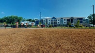 Located at 640 Lakeside Dr in Canton, Georgia, this outdoor dog park offers a generously sized, single-level open area designed specifically for pets and their owners. The ground is blanketed with a thick layer of wood chip mulch, providing a comfortable and low-maintenance surface suitable for dogs of all sizes to run and play freely.

The park is fully enclosed by a sturdy black wrought iron-style fence, ensuring the safety of all four-legged visitors. Decorative wooden pillar posts are spaced evenly along the perimeter, interspersed with neatly planted evergreen arborvitae trees that add vertical greenery and a defined, park-like aesthetic. Entry gates provide convenient access points throughout the enclosure.

The surrounding landscape is characterized by a dense canopy of mature trees, creating a natural, wooded backdrop that enhances the sense of seclusion and tranquility. The open sky above allows for abundant natural light throughout the day. Views of the adjacent residential community — a multi-story building with craftsman-style architectural detailing, board-and-batten siding, and balconies — are visible from within the park, situating the amenity within a well-planned residential setting. A paved walkway and bench areas along the perimeter offer comfortable resting spots for pet owners.