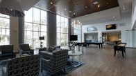 This sophisticated single-floor clubhouse offers an expansive, open-concept layout designed for both relaxation and social engagement. The space is anchored by dramatic floor-to-ceiling windows that flood the interior with abundant natural light while framing views of the outdoor surroundings, including palm trees and what appears to be a pool area beyond. The soaring ceiling, clad in warm wood paneling, creates an inviting contrast against the crisp white walls and adds a refined architectural character to the room. A statement chandelier with exposed bulbs serves as a central focal point, complementing the recessed lighting throughout. The lounge area is furnished with plush, contemporary seating — including a sectional sofa and patterned accent chairs — arranged around a sleek round coffee table atop a stylish area rug. Adjacent to the lounge, a billiards table provides a recreational centerpiece, while a wall-mounted flat-screen TV enhances the entertainment offerings. The hardwood flooring extends throughout, unifying the space with a warm, polished aesthetic. Further in the background, a dining or bar area is visible, suggesting multiple distinct zones within this well-appointed gathering space. The overall ambiance is modern, upscale, and welcoming.