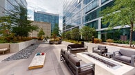 Located at 51 E 14th Street in Chicago's South Loop, this outdoor amenity terrace offers a well-appointed urban retreat set against the backdrop of the city's modern skyline. The terrace is thoughtfully divided into distinct zones, each designed for relaxation and recreation. A central lounge area features sleek, cushioned modular seating arranged around a built-in rectangular fire pit, creating an inviting gathering space for year-round enjoyment. Raised concrete planters filled with ornamental grasses, ferns, flowering shrubs, and mature shade trees introduce lush greenery and natural texture throughout the space. Smooth large-format pavers define the main walkways and seating areas, while decorative river rock beds add visual contrast and a refined, low-maintenance landscape aesthetic. A dedicated recreation zone includes cornhole boards and hanging egg chairs beneath shade sails, providing a casual, playful atmosphere. Softer seating vignettes with neutral-toned lounge chairs and benches offer quieter spots for relaxation amid the garden plantings. The terrace enjoys open sky views and is framed by the glass curtain walls of the surrounding contemporary high-rises, blending urban energy with tranquil outdoor living. Bollard lighting throughout ensures the space remains functional and atmospheric into the evening hours.