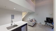 This contemporary two-floor loft-style space showcases a thoughtfully designed open-concept layout that blends modern finishes with functional living. On the main level, the kitchen is a standout feature, equipped with stainless steel appliances including a gas range, microwave, and refrigerator, complemented by dark espresso cabinetry, a brick-accented backsplash, and a generous white countertop island with an integrated sink. The kitchen flows seamlessly into the living area, which benefits from soaring double-height ceilings and a clerestory window that draws in natural light from above. A glass-panel door opens directly onto a private balcony, extending the living space outdoors. The main floor also includes a clean, well-appointed bathroom finished with large white tiles, a combination tub and shower, and dark wood vanity cabinetry. A staircase with white-painted steps leads to the upper level, where the bedroom features plush carpeting and an architecturally distinctive exposed ceiling with painted ductwork and piping — a hallmark of loft-style design. The upper level overlooks the main living area through an open railing, reinforcing the sense of vertical space. Polished concrete flooring on the main level and soft carpet upstairs create a pleasing contrast throughout this well-designed urban retreat.