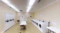 This communal laundry facility is a clean, efficiently organized single-floor space designed to accommodate the laundry needs of multiple users. The room features a prominent row of front-loading washing machines and dryers neatly aligned along one wall, offering ample capacity for simultaneous use. The machines are modern and well-maintained, with clearly visible control panels for ease of operation. A large folding table is centrally positioned, providing a convenient surface for sorting and folding laundry. Along the far wall, a utility sink offers additional functionality, accompanied by a drinking water fountain nearby. Informational notices and instructions are posted on the walls, ensuring users can operate the equipment with ease. The space is finished with warm, light-yellow walls accented by white crown molding, giving the room a clean and welcoming feel. Bright flush-mounted fluorescent ceiling panels ensure the entire room is well-lit. The flooring is smooth and easy to maintain, contributing to the overall tidy appearance. A single door provides access to the room, complementing the straightforward, practical layout. Overall, the space is thoughtfully designed to be functional, comfortable, and user-friendly for shared laundry use.