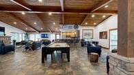 This single-story community amenity center is designed to provide residents with a wide range of functional and recreational facilities under one roof. The centerpiece is a spacious clubhouse featuring soaring vaulted ceilings with rich wood beam detailing and recessed lighting, creating a warm and inviting atmosphere. The open-concept layout accommodates a pool table, multiple seating arrangements with leather furniture, a wall-mounted flat-screen TV, and a dedicated computer workstation area. A built-in kitchen with warm wood cabinetry, stainless steel appliances, a mosaic tile backsplash, and a bar-height counter with seating provides a communal food and beverage area. The entrance is accented by striking stacked stone columns that add a rustic, lodge-like character. Natural light flows in through multiple windows throughout. The fitness center is equipped with treadmills, an elliptical, a rowing machine, and weight equipment, with motivational wall graphics and mirrored walls enhancing the workout environment. The indoor spa area features a tiled hot tub, lounge seating, and a wall-mounted TV for relaxation. A fully equipped laundry facility rounds out the amenities, offering stacked commercial washers and dryers for resident convenience.
