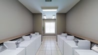 This single-floor laundry facility is designed with functionality and cleanliness in mind. The room features a symmetrical layout, with rows of white washers and dryers lining both sides of a central walkway, providing ample capacity for multiple users. Above each row, wooden-edged folding shelves offer a convenient surface for managing laundry. The flooring consists of light-colored ceramic tiles, contributing to a bright and easy-to-maintain environment. The walls are finished in a neutral taupe tone, lending a calm, understated aesthetic to the space. Overhead, recessed fluorescent light panels ensure thorough, even illumination throughout the room. A single window centered on the far wall, fitted with white horizontal blinds, allows natural light to supplement the artificial lighting while maintaining privacy. Ventilation is provided through ceiling-mounted HVAC vents. The overall design prioritizes a clean, orderly atmosphere, making the space practical and comfortable for everyday use.