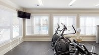 This fitness center is designed to support a full range of workout routines within a well-organized, single-level space. The interior features neutral beige walls, crown molding, and textured ceilings that lend a polished, finished appearance. Durable rubber flooring throughout provides both safety and practicality for exercise use.

The space is divided into distinct functional zones. The cardio area is equipped with multiple treadmills, elliptical machines, stationary bikes, and a recumbent cycle, all positioned near large windows fitted with horizontal blinds. These windows allow natural light to fill the space and offer views of an outdoor pool area. A wall-mounted flat-screen television is also present for entertainment during workouts.

The strength training area features a variety of cable and weight machines with red upholstered pads, along with adjustable benches and a dumbbell rack holding a full set of free weights. A medicine ball is also available for functional training exercises.

Entry to the fitness center is through elegant French doors with glass panes, contributing to the space's refined aesthetic. Overhead fluorescent lighting ensures consistent illumination throughout, complementing the natural light from the surrounding windows.