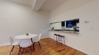 This single-floor residence is thoughtfully designed with a cohesive modern aesthetic throughout. Warm-toned hardwood flooring flows continuously across the open-plan living, dining, and kitchen areas, contributing to a sense of unity and spaciousness. The ceilings are notably tall, lending an airy, open quality to each room.

The kitchen is galley-style, featuring dark cabinetry, grey countertops, stainless steel appliances, a classic subway tile backsplash, and a distinctive penny-tile floor. A pass-through counter opens directly to the dining area, where a compact round dining table and modern chairs create an inviting casual space. Two leather barstools line the counter, offering an additional seating option.

The living area is comfortably arranged with a sofa, mid-century modern coffee table, and a flat-screen television. A sliding glass door leads to a private outdoor patio, allowing natural light to fill the room. The bedroom is calm and well-proportioned, featuring tall ceilings, soft neutral walls, and warm hardwood floors. The bathroom is finished with herringbone-pattern tile flooring, a dark vanity, and a tub-shower combination framed by white tile walls.