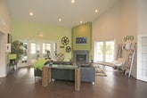 Located at 4401 Morris St NE in Albuquerque, New Mexico, this single-story community clubhouse offers a well-appointed gathering space designed for comfort and functionality. The interior is characterized by soaring vaulted ceilings that lend a sense of openness throughout the main areas, complemented by rich dark hardwood flooring and a neutral color palette with green accent walls.

The main lounge area serves as the heart of the space, anchored by a striking floor-to-ceiling stone fireplace with a wall-mounted flat-screen television above. French doors on multiple sides of the lounge flood the room with natural light and provide views of the surrounding landscaping. Adjacent to the lounge is a social gathering area featuring sleek chrome-and-black bar-height tables and stools, ideal for casual seating and conversation.

A bar counter with pendant lighting and a beverage vending machine adds convenient refreshment options for guests. A secondary lounge area offers additional seating with modern leather sofas and side tables, while a reading nook with magazine racks and shelving provides a quieter retreat. The fitness center, accessible through French doors, is equipped with cardio and strength-training equipment. Recessed lighting throughout ensures a well-lit environment across all areas of the clubhouse.