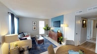 This thoughtfully designed single-floor residence offers a welcoming and contemporary living experience. The open-concept layout seamlessly connects the living and dining areas, creating a fluid and functional space ideal for both relaxing and entertaining. The living area is anchored by a striking teal accent wall that serves as a bold focal point, complemented by neutral gray walls that keep the overall palette balanced and modern. A wall-mounted flat-screen television is positioned above a warm, wood-toned media credenza, adding both style and practicality to the space. Light wood-style flooring runs throughout, enhancing the bright and airy atmosphere. A large area rug in deep navy blue grounds the seating arrangement, which features a plush white sofa accented with decorative throw pillows. Natural light filters in through a tall window, illuminating the space and highlighting the carefully chosen décor, including potted fiddle-leaf fig plants that bring a touch of greenery indoors. The dining area, visible in the foreground, includes a round wooden table with modern upholstered chairs, offering a cozy spot for meals. The textured ceiling adds a subtle architectural detail that completes the overall character of this well-appointed residence.