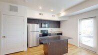 This thoughtfully designed one-bedroom, one-bathroom unit spans two floors, making smart use of every square foot to deliver a comfortable and functional living space. The main floor showcases a modern kitchen as its centerpiece, featuring rich dark-wood cabinetry extending to the ceiling for ample storage, a granite-topped island that doubles as a breakfast bar, stainless steel and black appliances including a refrigerator, electric range, microwave, and dishwasher, and a single-basin sink. Light wood-style flooring runs throughout the main living area, lending warmth and a sense of continuity. A clean, white hallway connects the main living spaces and leads to the full bathroom, which is finished with a dark-wood vanity, white countertop, a large mirror, and a tub-shower combination — all carried on the same light wood-style flooring. The bedroom is a cozy retreat carpeted in neutral tones, with a ceiling fan, and natural light filtering in through dual transom-style windows positioned near the ceiling. The second floor offers an additional carpeted room with a wall-mounted electrical panel, suitable for storage or utility purposes. Throughout the unit, the clean white walls, recessed lighting, and cohesive finishes create a bright, modern, and welcoming atmosphere.