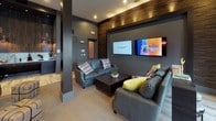 This single-floor amenity lounge is thoughtfully designed with a modern industrial aesthetic, combining dark gray walls, textured wood accent panels, and mixed flooring—including large-format marble-look tile and plush herringbone carpet—to create a dynamic and inviting atmosphere. The space is organized into several distinct zones that flow seamlessly into one another. A game area anchors one section of the room, featuring a rustic wood shuffleboard table set against a padded tile wainscoting wall adorned with framed black-and-white sports photography, adding a curated, club-like character. Adjacent to this is a fully equipped wet bar with a granite countertop, a kegerator, a glass-door beverage refrigerator, a microwave, and a metallic penny-tile backsplash illuminated by stylish gold directional sconces. The lounge area offers plush leather sofas centered around a coffee table, with wall-mounted flat-screen TVs and a textured wood feature wall above, creating an ideal space for relaxed gathering. Near large windows, a bistro-height round table with leather-topped stools provides a casual spot with views of the surrounding streetscape. The overall ambiance is sophisticated yet comfortable, with recessed lighting and layered design details throughout.