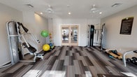 Located at 5712 IH-45 N in Wilmer, Texas, this single-floor fitness center is thoughtfully designed to support a comprehensive workout experience. The space is finished with a modern patchwork-style carpet in neutral tones, lending a clean and contemporary aesthetic throughout.

The cardio zone is positioned along a windowed wall, where multiple large windows fitted with blinds allow generous natural light to fill the room. Two treadmills and two elliptical machines are neatly arranged in this area, complemented by ceiling fans overhead that promote airflow and comfort during workouts.

Adjacent to the cardio area, a strength training section houses a multi-station cable machine and a pair of adjustable weight benches, providing options for a wide range of resistance exercises. A well-stocked dumbbell rack lines one wall, offering a full selection of free weights for users of varying fitness levels. A posted Fitness Center Rules sign indicates a managed and organized environment.

A dedicated accessories corner features a stability ball rack stocked with various sized exercise balls, along with additional floor space for stretching or functional training. Double glass French doors connect to an adjoining area, enhancing the sense of openness and allowing visual connectivity within the facility. Recessed lighting and ceiling fans are distributed throughout, ensuring the space remains well-lit and comfortable at all times.