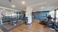 This fitness center offers a comprehensive workout facility thoughtfully organized into distinct training zones across a single open floor. The space is finished with durable wood-look flooring throughout, complemented by a clean white and deep navy-blue color palette that gives the interior a modern, energetic ambiance. Recessed ceiling lighting ensures the space is well-lit, while a ceiling fan provides additional air circulation in the strength training area.

The cardio zone lines one side of the facility and is equipped with a full array of TRUE-brand treadmills, elliptical trainers, upright bikes, and stair climbers. Large windows and a glass entry door flood this section with natural light and provide views of the surrounding outdoor area. A wall-mounted flat-screen TV is positioned for convenient viewing during workouts.

The free weights area features a dedicated rubber-matted platform with adjustable benches, a full dumbbell rack, and a multi-station power rack, all reflected by a floor-to-ceiling mirror that amplifies the sense of space. Adjacent to this zone, a selection of Hoist selectorized strength machines caters to targeted muscle training. A structural column divides the space naturally, creating defined workout areas without compromising the open, airy feel of the overall facility.