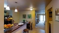 This single-floor residence offers a cohesive and functional open-concept layout designed for comfortable everyday living. The space is anchored by a kitchen bar counter with a dark granite-style surface, ideal for casual dining or entertaining. A pendant light hangs above the bar, adding a warm, focused glow, while a flush-mount ceiling fixture illuminates the surrounding area. Adjacent to the bar, a cozy dining nook accommodates a small round table with chairs, creating an intimate setting for meals. The walls are painted in a warm yellow tone, contributing to an inviting and cheerful ambiance throughout the common areas. Decorative tree artwork adorns the walls, adding a touch of character to the space. The flooring transitions from tile in the kitchen and dining area to soft carpet as the space flows toward the living room. The living room is bright and airy, with natural light streaming in through large windows or glass doors at the far end, offering a welcoming retreat. The overall design balances practicality with warmth, making it a comfortable and well-appointed single-level home.