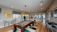 This single-floor recreational space in Houston, TX (77049) is thoughtfully designed to serve as a lively gathering and entertainment hub. The open layout is anchored by warm hardwood-style flooring that runs throughout, complemented by classic wainscoting along the lower walls and recessed lighting that keeps the interior bright and welcoming.

At the center of the room, a full-sized pool table rests atop a bold geometric area rug in teal, yellow, black, and white, with a pendant light fixture hanging directly overhead to illuminate gameplay. Nearby, vintage arcade cabinets add a nostalgic touch and serve as eye-catching focal points.

Seating is varied and plentiful. High-top tables with bar-height stools are positioned near the windows, offering casual spots for conversation or casual dining. A round dining-height table surrounded by modern white chairs provides additional gathering space. A wall-mounted bar counter with sleek gray stools lines one side of the room for more relaxed seating.

Natural light flows in generously through multiple windows dressed with patterned Roman shades and slatted blinds. Large glass French doors connect to an adjacent fitness area, enhancing the sense of openness. Colorful wall art and indoor greenery add personality, making this space feel energetic yet comfortable.