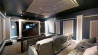 This dedicated home theater is a masterfully designed entertainment space that combines refined architectural detail with high-performance functionality. The room's most striking feature is its ornate coffered ceiling, which showcases a central oval medallion framed by decorative molding and beadboard paneling — a classical touch that elevates the space well beyond a typical media room.

The walls are finished in a deep, rich blue-gray tone that enhances the cinematic atmosphere and minimizes light reflection during screenings. Framed acoustic panels are symmetrically mounted along the side walls, ensuring optimal sound absorption and audio clarity throughout the room. White decorative columns flank the viewing area, adding an architectural grandeur that reinforces the theater's upscale character.

Track lighting installed along the ceiling perimeter provides adjustable ambient illumination, while a large flat-screen display serves as the visual focal point, mounted within a custom dark wood entertainment surround. Seating is arranged in two tiered rows of plush, oversized reclining chairs in a warm gray upholstery, each designed for comfort during extended viewing. A patterned area rug anchors the seating arrangement, adding warmth and texture to the floor. Together, these elements create an immersive, cinema-quality experience within a beautifully appointed private setting.