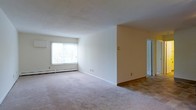 This single-floor space presents a clean, open living area finished in neutral tones throughout. The main room features wall-to-wall carpet in a warm beige hue, contributing to a comfortable and understated atmosphere. A large window along one wall allows natural light to fill the space, creating a bright and airy feel. Below the window, a baseboard heating unit runs the length of the wall, providing efficient climate control. The walls are painted in a soft, light cream tone that complements the flooring and enhances the sense of openness. A partial wall divides the main living area from an adjoining space, which transitions to hard surface flooring — likely tile or laminate — and leads toward what appears to be an additional room or hallway visible through a doorway. The layout is straightforward and functional, with clean lines and minimal architectural detailing. The overall ambiance is calm and neutral, offering a versatile blank canvas well-suited to a variety of interior arrangements and personal styles.