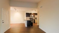 This single-floor space showcases a contemporary open-concept design that seamlessly integrates the living area and kitchen. The rich, dark hardwood flooring extends throughout, providing a warm and cohesive aesthetic. Elevated ceilings add a sense of spaciousness, while track lighting installed along the ceiling delivers both functional illumination and a modern touch.

The kitchen is a standout feature, outfitted with dark espresso cabinetry that contrasts sharply against light-toned walls. A peninsula with a dark countertop serves as both a workspace and a casual dining or entertaining area. Stainless steel appliances — including a gas range, over-the-range microwave, and a French door refrigerator with an ice and water dispenser — complete the kitchen's modern, functional profile. A stainless steel sink is integrated into the peninsula.

The open living area adjacent to the kitchen is bright and versatile, with clean white walls and additional track lighting, offering ample flexibility for furniture arrangement. The overall ambiance is sleek and minimalist, with a neutral color palette that creates a clean, contemporary atmosphere throughout the space.