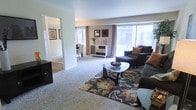 The Bavarian is a well-appointed 2-bedroom, 1-bathroom single-story residence that makes efficient use of its generous square footage. The living room serves as the heart of the home, featuring plush wall-to-wall carpeting, neutral-toned walls, and large sliding glass doors that flood the space with natural light while providing access to an outdoor area. The open layout allows for versatile furniture arrangements, as evidenced by the ample room for a full seating area and entertainment setup. A decorative area rug anchors the living space, adding warmth and visual interest. Adjacent to the living room, the dining area is visible through an open passageway, featuring hard flooring and space for a full dining set, making it ideal for everyday meals or entertaining. The transition between rooms flows naturally, creating a cohesive and connected feel throughout the home. Additional wall space throughout accommodates artwork and personal touches. The combination of carpeted living areas, hard-surface dining space, and abundant window placement gives this residence a bright, welcoming ambiance suitable for comfortable day-to-day living.