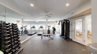 This fitness center offers a comprehensive workout environment spread across a single, open floor. The space is designed with functionality and comfort in mind, featuring durable rubber flooring throughout that provides both safety and shock absorption during exercise. A large floor-to-ceiling mirror runs along one wall, visually expanding the space and allowing users to monitor their form during workouts.

Natural light filters in through multiple windows, creating a bright and energizing atmosphere, while recessed ceiling lights and a ceiling fan ensure consistent illumination and airflow. The cardio section is well-appointed with treadmills, an elliptical trainer, and a stationary recumbent bike, all positioned to take advantage of the natural light from the windows. A multi-function cable machine provides a wide range of strength training options, complemented by a dedicated free weights area featuring a full dumbbell rack, exercise bench, stability balls, and additional resistance training equipment.

The layout is thoughtfully organized, with open floor space in the center allowing for stretching or bodyweight exercises. The neutral color palette of whites and soft grays contributes to a clean, modern aesthetic. Entry is provided through a paneled exterior door with a window, and the space also connects to an adjacent utility area. Overall, this fitness center is a practical and well-maintained facility suited for a full range of workout routines.