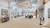 Located at 100 Fox Meadow Drive in Moorestown, NJ, this single-floor community amenity facility offers a generously proportioned, thoughtfully designed interior spanning five distinct areas. The centerpiece is a sweeping great room with soaring ceilings, geometric coffered detailing, and elegant chandelier lighting that creates a bright, welcoming atmosphere. Natural light pours in through large windows, offering views of the surrounding landscape. Seating areas are arranged throughout, anchored by a linear fireplace with marble surround and built-in wood shelving. Adjacent to the main lounge is a dedicated screening and media room, furnished with tiered rows of plush gray recliners and wall sconce lighting for a true theater experience. A large-screen TV lounge provides additional viewing space with comfortable seating. The game room is a standout feature, housing a full-size billiards table beneath a statement pendant light, high-top seating, and direct outdoor access, all complemented by a striking geometric accent wall. A welcoming entry vestibule with glass double doors and a smaller sitting lounge with colorful artwork and garden views round out the space. Throughout, neutral carpet, warm wood accents, and curated art create a cohesive, upscale ambiance ideal for community gathering and recreation.
