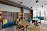 This thoughtfully designed single-floor communal space blends contemporary aesthetics with functional versatility. The layout is divided into distinct zones, each offering a different social or work-focused experience. A welcoming entrance area features polished concrete flooring, a striking wood-slat accent wall housing a mounted display screen, and smooth cylindrical columns that anchor the open layout.

The lounge area is anchored by a bold teal sectional sofa positioned beneath floor-to-ceiling windows, flooding the space with natural light and offering expansive outdoor views. A leather sofa nearby echoes the warm, mid-century modern palette. Built-in banquette seating with tall striped upholstered panels and teal velvet cushions, accented by yellow throw pillows and warm Edison-style wall sconces, creates intimate dining nooks complemented by round wood tables and mid-century chairs.

A vibrant teal feature wall adorned with three-dimensional white hexagonal art installations adds a sculptural focal point. Private meeting rooms enclosed by black-framed glass walls offer a more focused setting, one featuring bold botanical wallpaper. A compact, high-backed acoustic pod provides an additional private nook. Geometric-patterned carpet tiles and wood-look flooring define transitions between zones, while dual elevators with wood-panel surrounds complete this well-appointed space.