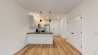 This thoughtfully designed single-floor unit offers a clean, modern aesthetic with light hardwood flooring and crisp white walls throughout. The open-concept kitchen and living area serves as the heart of the space, anchored by a peninsula with a dark countertop and seating potential. The kitchen is well-appointed with two-tone cabinetry — white upper cabinets paired with slate-blue lower drawers — complemented by a light subway tile backsplash, stainless steel appliances including a refrigerator, range, and microwave, and pendant lighting that adds a stylish focal point above the peninsula. Recessed lighting and a clear sightline to the entry door enhance the airy, open feel of the main living space. The bedrooms are bright and generously proportioned, each featuring light hardwood flooring, neutral walls, and windows with views of surrounding greenery. Select bedrooms include built-in closet storage with sleek panel doors and in-unit climate control units. Two bathrooms serve the space: one featuring large-format gray tile flooring, a tub-shower combination with glass enclosure, a white vanity, and a mirror; the other offering a corner glass-enclosed walk-in shower, a white vanity, and coordinating tile finishes. Together, these spaces deliver a cohesive, contemporary living environment.