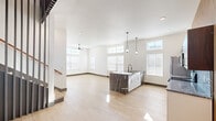 This modern two-story home on Village Run Parkway in Little Rock, Arkansas, offers a thoughtfully designed layout across two floors. The main level features an open-concept living and kitchen area bathed in natural light from an abundance of windows, including transom windows that draw in additional light and add architectural interest. Light hardwood flooring runs throughout, complementing the crisp white walls and dark trim accents. The kitchen is a standout, featuring white cabinetry, dramatic dark granite countertops with a waterfall island, stainless steel appliances, and stylish pendant lighting. The adjacent living room includes a built-in fireplace and ceiling fan. A staircase with a contemporary metal railing leads to the upper level, where both bedrooms are located. The spacious primary bedroom boasts soaring vaulted ceilings with exposed beams, a ceiling fan, and ample natural light. The second bedroom also features elevated ceilings, transom windows, and a bright, open feel. The three bathrooms are finished with warm wood vanities, vessel sinks, granite countertops, and modern fixtures. The home's clean, contemporary aesthetic is carried consistently throughout every room, creating a cohesive and polished living environment.