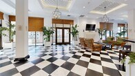 Located at 1026 S Broadway in the heart of Los Angeles, this sophisticated single-floor space offers a generously scaled interior defined by striking architectural details and a refined, classic aesthetic. The most commanding feature is the bold black-and-white checkered tile flooring that stretches across the entire space, lending a timeless, grand hotel ambiance throughout.

Elevated coffered ceilings with intricate latticed inlays and ornate crown molding add significant visual depth, while a series of wrought-iron chandelier pendants cast warm, layered lighting across the open floor plan. Bamboo Roman shades frame large windows and glass-paned entry doors on multiple walls, allowing generous natural light to filter in while maintaining a warm, curated atmosphere.

Multiple lounge seating arrangements are distributed throughout, featuring green velvet sofas, rattan armchairs, and glass-topped coffee tables accented by lush tropical plants. A bar counter area with dark cabinetry and bar stools anchors one section of the space, complemented by a white fireplace mantle with a mounted television nearby. An arched doorway leads to an additional service or kitchen area. Curated framed artwork adorns the white walls, completing the polished, hospitality-inspired character of this distinctive Los Angeles space.
