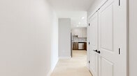 This thoughtfully designed single-floor residence combines clean modern aesthetics with functional living spaces. Light hardwood flooring runs continuously throughout the unit, creating a seamless, airy feel. A hallway leads from the entry into the heart of the home, where the kitchen serves as a standout feature. The kitchen is outfitted with warm wood-toned cabinetry, white quartz countertops, a mosaic tile backsplash, stainless steel appliances, and a peninsula that offers additional prep and seating space. Recessed lighting ensures the kitchen is well-lit at all times. The bedrooms are generously proportioned and bathed in natural light, thanks to large black-framed windows that frame urban views of the surrounding cityscape and autumn foliage. These bold architectural windows add a striking contemporary contrast to the neutral gray and white walls. The bathroom is finished with dark, high-gloss floor tiles, a classic white subway tile surround encasing a full soaking tub, a floating wood-toned vanity, white countertop, and a circular mirror with modern wall-mounted lighting. Throughout the space, clean white trim, panel-style doors with matte black hardware, and a neutral color palette contribute to a cohesive and refined modern aesthetic.