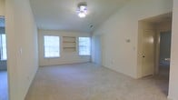 This single-story space centers around a spacious main living area defined by vaulted ceilings that lend an airy, open feel to the room. The ceiling is equipped with a multi-light ceiling fan, providing both lighting and ventilation. Neutral-toned carpet covers the floor throughout, creating a clean and cohesive look. Two large windows along the far wall allow abundant natural light to fill the space, offering views of the surrounding greenery outside. Between the windows, built-in shelving units provide functional display and storage space, adding architectural character to the room. A vertical blind-covered door or window opening sits to the right of the built-ins, offering additional light control. The walls are finished in a soft, neutral tone that complements the overall brightness of the space. A wall-mounted control panel — likely for climate or security — is visible on the right-hand wall, indicating modern conveniences are in place. The layout is open and flexible, allowing for a variety of furniture arrangements to suit different needs. Overall, the space presents a comfortable, well-lit environment with practical built-in features.