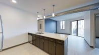 This single-floor space showcases a thoughtfully designed open-concept layout that seamlessly connects the kitchen and living areas. The kitchen is anchored by a sleek, extended island with a light-colored countertop and an integrated sink, complemented by dark espresso cabinetry with contemporary hardware. Stainless steel appliances add a polished finish to the space. Overhead, cylindrical pendant lights hang above the island, while an exposed metallic ductwork element running along the ceiling lends an industrial character to the design. The open living area beyond features a striking blue accent wall that creates a visual focal point, contrasting with the neutral white walls and light-toned flooring throughout. Natural light filters in through windows and a glass door that leads to an outdoor area, brightening the interior and enhancing the sense of openness. The clean lines, modern fixtures, and carefully chosen color palette come together to create a space that is both functional and stylish, offering a contemporary urban living experience with a distinct industrial edge.