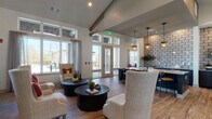 The clubhouse at 575 Concord Rd in Jefferson, Georgia, is a thoughtfully designed single-floor community space that seamlessly blends modern aesthetics with comfortable, functional living. The interior is defined by soaring vaulted ceilings accented with exposed wooden beams, creating an open and airy atmosphere throughout. Rich wood-look flooring runs continuously across the main areas, anchoring the space with warmth and visual cohesion.

The expansive lounge area is arranged into multiple distinct seating zones, offering a variety of settings for both relaxed conversation and casual gatherings. Plush high-back chairs, neutral sofas, and leather accent seating are complemented by dark wood round coffee tables and carefully curated décor. A large flat-screen TV is built into a teal-toned accent wall flanked by built-in shelving, serving as the focal point of one lounge area.

The kitchen features a generous central island with a light quartz countertop, dark cabinetry, and stainless steel appliances. Pendant lantern-style lights hang overhead, adding an industrial-chic touch. A full prep and service counter runs along one wall, finished with a tile backsplash and equipped with a sink and ample storage.

Abundant natural light pours in through oversized windows and glass-paneled doors, offering views of the surrounding outdoor areas. The overall ambiance is modern, welcoming, and well-appointed — ideal for community events and everyday resident use.