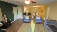 This single-floor residence offers a thoughtfully designed open-concept layout that seamlessly connects the main living areas. Rich hardwood flooring flows throughout, anchoring the warm, inviting ambiance created by golden-yellow walls and bold accent colors, including a striking dark green feature wall near the entryway.

The living area is centered around a stylish area rug with a geometric pattern, complemented by two blue accent chairs and a glass-top coffee table. A ceiling fan overhead adds both function and charm to the space.

The dining area sits adjacent to the living room, featuring a sleek glass dining table with white chairs and tasteful wall art, creating an elegant yet casual setting for meals and gatherings.

The kitchen is open to the main living space and features white cabinetry, modern appliances including a refrigerator, and a raised breakfast bar with black bar stools — ideal for casual dining or entertaining. Pendant lighting above the bar adds a polished finishing touch.

The layout is practical and cohesive, with clear sightlines across all three zones, making the space feel open and connected. The combination of warm tones, contemporary furnishings, and quality finishes gives this residence a comfortable and stylish character throughout.