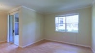 This single-floor space presents a clean, understated interior with a straightforward and functional layout. The room showcased in the image features light hardwood flooring that runs throughout, contributing a warm, neutral foundation to the space. The walls are finished in a soft beige tone, complemented by crisp white crown molding along the ceiling perimeter, adding a subtle architectural detail that elevates the overall aesthetic.

A large, multi-pane window is a standout feature of the room, allowing a generous amount of natural light to fill the interior. The window offers views of the surrounding outdoor environment, including lush green shrubbery and neighboring buildings, providing a connection to the outside while maintaining the brightness of the space. White baseboards frame the perimeter of the room, maintaining a cohesive and tidy finish throughout.

The space is currently unfurnished, making it easy to assess its proportions and versatility. The overall ambiance is calm and neutral, offering a blank canvas well-suited for a variety of uses. The combination of natural light, warm flooring, and classic molding details creates a balanced and welcoming environment.