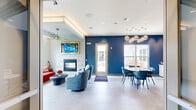 Located at 461 Railroad Ave in Westbury, NY, this thoughtfully designed community lounge at The Cornerstone Westbury offers a sophisticated and welcoming atmosphere across a single level. The space is entered through wide glass double doors that open into a bright, open-plan layout combining a living room and dining area, connected by a welcoming hallway.

The living area is anchored by a striking marble-surround fireplace topped with a gold-framed wall-mounted TV and complemented by a tray ceiling with warm cove lighting and a sculptural brass ceiling fan. Generous windows allow abundant natural light and outdoor views of lush greenery. Seating includes a plush red velvet sofa, patterned blue swivel chairs, and a sleek oval coffee table, creating a vibrant and eclectic aesthetic.

The dining zone features bold navy blue accent walls, a round white dining table with teal velvet chairs, and an eye-catching cluster of globe pendant lights in brushed gold. Additional casual seating is also available nearby.

A built-in refreshment bar serves as a functional highlight, complete with marble-look backsplash, a mirrored decorative wall panel, wood cabinetry, stainless steel under-counter refrigerators, a gold faucet, and a coffee station. Large-format light tile flooring flows throughout, unifying the entire space with a clean, modern finish.