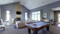 Located at 3520 Red Hook Xing in Columbus, Ohio, this single-floor community space is designed to impress with a thoughtfully appointed great room that serves as its centerpiece. The vaulted ceiling rises dramatically overhead, enhanced by a ceiling fan and recessed lighting that keep the space bright and comfortable. A floor-to-ceiling stacked stone fireplace anchors one wall, topped with a flat-screen television and framed by a clean white mantel, creating a striking focal point. Soft blue-gray walls lend the room a calm, sophisticated atmosphere, complemented by plush neutral-toned seating arranged for both conversation and relaxation. A full-sized billiards table occupies the center of the room, making it an ideal gathering space for recreation. Large multi-pane windows along the far wall flood the interior with natural light while offering views of the surrounding greenery, framed by elegant drapery panels. Decorative artwork adorns the walls, adding a refined, curated touch to the overall aesthetic. The carpeted flooring ties the space together, providing warmth and comfort underfoot. The combination of soaring architecture, recreational amenities, and inviting design elements makes this a versatile and welcoming communal destination.