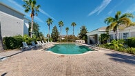 Located at 5337 Esplanade Park Cir in Orlando, Florida, this outdoor community amenity area offers a resort-inspired poolside experience within a lushly landscaped setting. The centerpiece is a generously sized freeform swimming pool with a curved, organic shape and striking turquoise water, complemented by a shallow wading ledge entry that provides easy and gradual access. The pool deck is finished with classic brick pavers that extend across the entire surround, creating a warm, durable surface that accommodates a full lineup of white lounge chairs — ideal for sunbathing beneath the Florida sun. Tall, mature palm trees are planted throughout the perimeter, providing both shade and a distinctly tropical atmosphere, while well-maintained hedges and lush tropical foliage serve as natural privacy borders around the space. A dedicated pool house structure is positioned at the edge of the deck, featuring bold blue French doors with glass pane detailing, tile flooring, and access to restroom facilities — adding both convenience and a pop of color to the overall aesthetic. Additional seating and a bar-style counter area are visible along the far perimeter, offering a social gathering spot. The open-air setting benefits from abundant natural sunlight and open sky views, reinforcing the relaxed, vacation-like ambiance of this well-appointed outdoor community space.