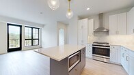 This thoughtfully designed 2-bedroom, 2-bathroom residence is laid out across a single floor, offering an efficient and functional floor plan. The open-concept kitchen and dining area serves as the heart of the home, featuring a central island with a light countertop, taupe cabinetry accented with gold hardware, and a pair of elegant glass globe pendant lights overhead. The full kitchen is outfitted with white shaker cabinets, white subway tile backsplash, quartz countertops, stainless steel appliances, and a gold-toned faucet — a clean, contemporary aesthetic throughout. Large black-framed windows flood the living and dining spaces with natural light and provide views of the surrounding urban environment. The primary bedroom is generously sized, complete with soft carpeting, a ceiling fan, a closet, and a large window. The second bedroom also features light hardwood-style flooring and a bright, airy feel. Both bathrooms are finished with warm wood-tone LVP flooring, white vanities with grey countertops, and matte black hardware and fixtures. The primary bath includes a glass-enclosed tub and shower with marble-style tile surround. Two hallways connect the rooms seamlessly, contributing to a smooth, well-organized flow throughout the residence.