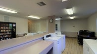 The laundry facility at Sugar Mill Creek is a single-floor, dedicated utility space designed with functionality and convenience in mind. The room is well-lit by a combination of fluorescent ceiling fixtures and natural light filtering through a windowed door, creating a bright and practical environment. Multiple commercial-grade washing machines are neatly arranged in a row, complemented by additional dryers to accommodate high-volume use. Generous laminate folding counters run along the perimeter, providing ample workspace for residents to sort and fold laundry with ease. The flooring consists of neutral ceramic tile, which is both durable and easy to maintain. Walls are finished in a soft neutral tone, contributing to a clean and orderly atmosphere. A wall clock is centrally positioned, keeping the space practical and user-friendly. Two doorways provide access points, enhancing the flow and accessibility of the facility. The overall layout is open and uncluttered, ensuring comfortable movement even when the room is in full use. This well-maintained laundry facility reflects a commitment to providing residents of Sugar Mill Creek with a reliable and efficient shared amenity.