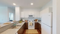 This well-designed single-floor residence offers a cohesive blend of modern finishes and functional layout. The kitchen is configured in a U-shape, featuring two-tone cabinetry with white upper cabinets and warm walnut-toned lower cabinets, granite-look countertops, a stainless steel sink, and a full suite of white appliances including a range, over-the-range microwave, and refrigerator. A breakfast bar with seating extends from the kitchen, providing a casual dining option. The living area is bright and open, benefiting from multiple windows that invite ample natural light and outdoor views. A ceiling fan adds comfort to the space, which features rich hardwood-style flooring carried throughout the unit. The bedrooms are generously sized, each with large windows, ceiling fans, and hardwood-style floors. One bedroom includes access to a spacious walk-in closet with wire shelving and hanging rods. The bathroom is elegantly appointed with a wood-tone vanity, granite-look countertops, a large mirror, and hardwood-style flooring. Clean white walls and recessed lighting throughout the unit contribute to a bright, contemporary ambiance that feels both airy and inviting.