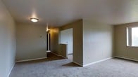This single-floor space showcases a straightforward, functional layout with a clean and neutral aesthetic throughout. The main living area is finished with wall-to-wall carpeting in a light tone, providing a comfortable underfoot feel, while the walls are painted in a warm greige hue that lends a cohesive, understated look to the interior.

A flush-mount ceiling light provides overhead illumination, keeping the space well-lit. The open design flows naturally toward a hallway corridor, where a partial wall and architectural cutout create a subtle visual divide between the living area and the adjoining spaces. The hallway, visible in the background, features darker flooring and leads to additional rooms, hinting at a thoughtfully segmented layout.

White baseboards run consistently throughout, adding a crisp contrast to the neutral walls and flooring. Electrical outlets and light switch plates are positioned at standard heights along the walls. The overall ambiance is calm and move-in ready, offering a blank canvas well-suited for a variety of furnishing and décor styles.