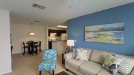 This single-floor space is designed around an open-concept layout that connects the living, dining, and kitchen areas into one cohesive, functional environment. The living area serves as the focal point, anchored by a bold blue accent wall adorned with a landscape painting that introduces a vibrant splash of color against the neutral beige walls and light carpeted flooring. A comfortable neutral sofa paired with a colorful patterned accent chair creates an inviting seating arrangement, while a floor lamp provides warm ambient lighting.

Flowing naturally from the living area, the dining space accommodates a dark-stained wooden table and chairs beneath a pendant light fixture, creating a dedicated area for meals and gatherings. The kitchen is positioned directly behind, featuring dark cabinetry, a peninsula counter that separates it from the dining area, and standard appliances including a refrigerator. Recessed and fluorescent ceiling lighting illuminate the kitchen efficiently.

Throughout the space, the open layout promotes a sense of airiness and ease of movement, making it well-suited for both everyday living and entertaining. The cohesive color palette and consistent flooring tie the distinct functional zones together harmoniously.