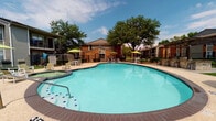 This outdoor amenity space is centered around a generously sized freeform swimming pool with crystal-clear turquoise water, complemented by an adjacent hot tub for year-round relaxation. The pool deck is finished in textured stamped concrete, providing both durability and visual appeal, and is furnished with multiple lounge chairs and side tables positioned for sunbathing and leisure. Bright green umbrella-shaded seating areas are arranged poolside, offering shaded spots to unwind. A standout feature of the space is a handsome wooden pergola sheltering a stone-clad outdoor bar area, creating an ideal gathering spot for residents. Lush, manicured green lawn surrounds portions of the deck, enhancing the resort-like atmosphere. Mature shade trees frame the perimeter, providing natural canopy and a sense of privacy. The surrounding buildings feature classic brick and siding exteriors with black wrought-iron fencing enclosing the pool area for safety and definition. A dedicated sunning patio adjacent to the main building offers additional lounge seating on stamped concrete. The overall ambiance is vibrant, inviting, and well-maintained, blending functional outdoor living amenities with attractive landscaping under open Texas skies.