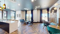 This thoughtfully designed single-floor communal space blends bold aesthetics with functional versatility. The lounge area greets visitors with striking dark teal ceilings, floor-to-ceiling navy drapery, and an abundance of natural light streaming through large windows that offer open outdoor views. Mid-century modern seating arrangements — including slatted armchairs and a central coffee table — invite relaxed conversation, while the distinctive chevron-patterned parquet flooring ties the entire space together with warmth and visual interest.

Adjacent to the lounge is a fully equipped kitchen featuring a dramatic floor-to-ceiling backsplash of glossy teal herringbone tiles that serves as the room's focal point. Dark cabinetry, white countertops, a stainless steel dishwasher, a built-in beverage cooler, and industrial-style bar seating make this kitchen ideal for both casual use and entertaining. Open wooden shelves add a decorative yet practical touch.

A dedicated dining nook rounds out the space with an elegant arched banquette wall adorned with botanical wallpaper and warm golden upholstery. Round wooden tables paired with plush emerald velvet chairs sit beneath sculptural brass sputnik chandeliers, creating an intimate and sophisticated dining atmosphere. The seamless flow between each zone makes this a highly functional and visually cohesive gathering space.