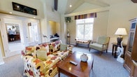 This single-floor space centers around a bright and inviting living area distinguished by soaring vaulted ceilings that lend a sense of openness and grandeur. The architectural design incorporates recessed lighting set into the pitched ceiling, complementing the abundant natural light that streams through a large, multi-pane window adorned with striped valances in warm earth tones. The window frames views of lush greenery, creating a pleasant connection to the outdoors.

The living area is furnished with a bold floral-patterned sofa in shades of red, orange, yellow, and brown, paired with coordinating upholstered seating and classic wooden accent chairs. A polished wood coffee table anchors the seating arrangement, topped with a ceramic vase for a decorative touch. Neutral carpet and light-toned walls provide a balanced backdrop to the colorful furnishings.

A recessed nook adjacent to the main living space features a built-in counter area, suitable for a small workspace or display area. A doorway leads to what appears to be an additional room or entry point, adding to the functional flow of the layout. Overall, the space exudes a cozy, well-appointed character with traditional design sensibilities throughout.