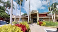 This single-story building presents a striking Florida Mediterranean architectural style, characterized by a warm stucco exterior in earthy tan and terracotta tones complemented by a classic clay tile roofline. The structure features a prominent covered entry portico, flanked by traditional wall-mounted lantern-style light fixtures that add an inviting, refined touch to the facade.

The approach to the building is defined by a decorative paver walkway that draws the eye directly toward a set of elegant glass double doors, creating a clear and welcoming sense of arrival. Towering royal palms line the entryway, rising dramatically above the roofline and contributing to the distinctly tropical Florida atmosphere. The surrounding landscaping is lush and meticulously maintained, featuring bold bursts of color from red and yellow tropical foliage, bromeliads, and ornamental shrubs that frame the building on all sides.

The exterior color palette — combining warm beige with accents of deep red — adds visual depth and character to the overall composition. Large windows punctuate the facade, suggesting well-lit interior spaces. The combination of architectural detail, curated tropical plantings, and the natural backdrop of a bright Florida sky creates an overall ambiance that is polished, inviting, and distinctly regional in character.