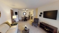 This single-floor residence offers a thoughtfully designed layout that balances comfort and functionality across its key living areas. The living room serves as the heart of the home, featuring plush carpeting, a large sliding glass door with vertical blinds that invites natural light, and ample space for relaxed seating arrangements. A ceiling fan overhead adds to the room's comfort. Adjacent to the living area, a dedicated dining space accommodates a round table and chairs beneath its own ceiling fan, with a charming round mirror adding a decorative touch to the wall nearby. Two interior doors provide convenient access to closets or storage. The galley-style kitchen is efficiently organized with light wood cabinetry, a double basin sink, a dishwasher, electric range, refrigerator, and a microwave, all set against tile flooring and sage-toned walls. The primary bedroom is generously sized, carpeted, and features a ceiling fan, a window with a view of lush greenery, and a closet. A versatile second room with soft blue walls offers flexible use as a home office or guest space. The bathroom is bright and clean, featuring a vanity with under-cabinet storage, a large mirror, and well-appointed fixtures. Throughout the residence, warm neutral tones and carpet flooring create a cohesive, welcoming ambiance.