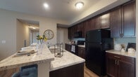 Located at 1 Kerry Park Cir in Delaware, Ohio, this single-floor residence offers a thoughtfully designed layout with modern finishes throughout. The kitchen is a standout feature, showcasing rich dark-espresso cabinetry extending to the ceiling, light granite countertops, and black appliances including a refrigerator, range, and built-in microwave. A spacious island with bar seating and an undermount sink serves as both a functional workspace and a casual dining area, all set atop durable hardwood-style flooring. The open layout connects the kitchen seamlessly to the living area, which is carpeted and bathed in natural light from large sliding glass doors fitted with vertical blinds. The bedrooms are comfortably sized with neutral carpet, natural light from windows with blinds, and generous closet space — including a walk-in closet with built-in shelving and rod storage. The bathroom features a glass-enclosed walk-in shower, a dark-vanity with granite countertop, and tile flooring. Warm, neutral tones are consistent throughout the residence, creating a cohesive and inviting ambiance. The overall design balances practicality and comfort, making this a well-equipped home in Delaware, Ohio.