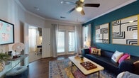 This single-story residence showcases a thoughtfully designed interior highlighted by bold color choices and eclectic styling throughout. The living room serves as a central gathering space, featuring rich hardwood-style flooring, crown molding, a ceiling fan with integrated lighting, and a dramatic teal accent wall adorned with geometric artwork. French doors invite natural light and provide outdoor access, while large windows dressed with layered curtains further brighten the space.

The dining area continues the teal accent wall theme, complemented by an eclectic gallery wall arrangement and warm wood flooring. The primary bedroom features a striking dark accent wall, carpeted flooring, and access to an adjoining room. A secondary bedroom carries a softer aesthetic with a muted blue-green accent wall, carpet, and generous natural light from a large window.

The bathroom offers hardwood-style flooring, a combination tub and shower with white tile surround, a single vanity with wood cabinetry and stone-look countertops, and a large mirror. A dedicated built-in workspace provides cabinetry and countertop surface with its own separate entry. Together, these spaces create a cohesive, design-forward interior with a vibrant, eclectic character throughout.