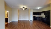 Vineland Carriage Homes presents a thoughtfully designed single-floor residence that combines comfort and functionality in an open, welcoming layout. The living area is generously sized, featuring warm hardwood-style flooring that extends seamlessly throughout the space, creating a cohesive and inviting atmosphere. A ceiling fan with a light fixture anchors the room, providing both airflow and ambient lighting, while soft, neutral-toned walls enhance the bright and airy feel of the interior.

The kitchen flows directly from the living area in an open-concept arrangement, making the space ideal for both everyday living and entertaining. It is outfitted with rich, dark espresso cabinetry that offers ample storage, complemented by granite-style countertops and black appliances, including a range and over-the-range microwave. The contrast between the dark cabinetry and the lighter walls and flooring gives the kitchen a bold, modern aesthetic.

The overall design of the home emphasizes clean lines, practical finishes, and a warm, neutral palette. The single-floor layout ensures easy accessibility and a smooth flow between spaces. Vineland Carriage Homes delivers a well-appointed residence with stylish, durable finishes suited for comfortable everyday living.